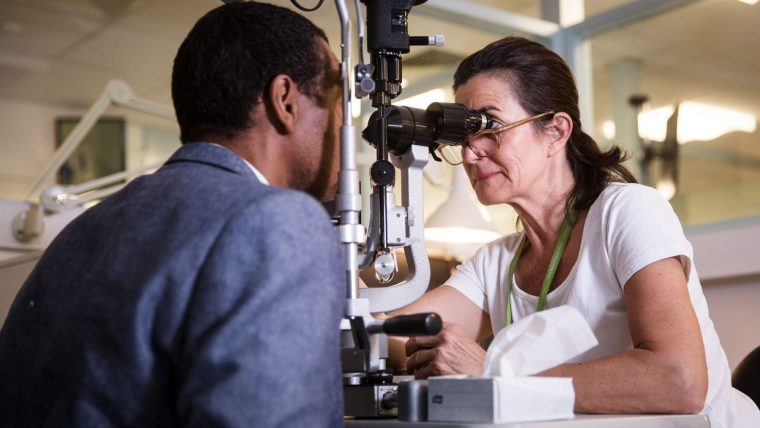 Patient having an eye exam. He is resting his head the support whilst a clinician looks into his eyes through a microscope.