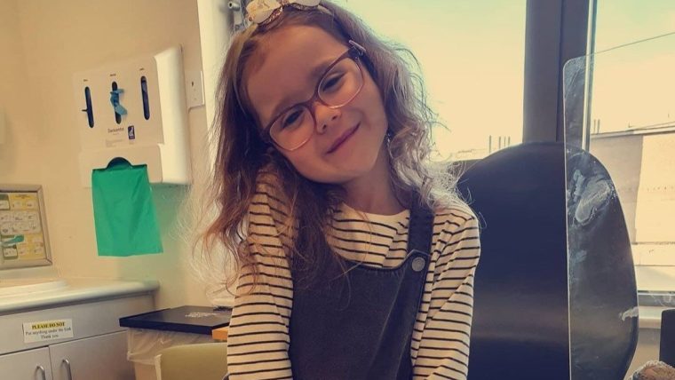 A young girl sitting in a doctors office and smiling. She is wearing glasses, a white bow headband and a white stripy top.