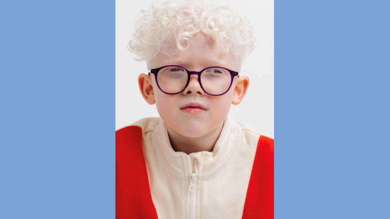 A professional photo of Milo, an eight year old boy with albinism. He is wearing a red and white jumper, and dark round glasses.