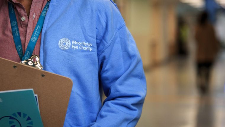 A close up of someone wearing a blue Moorfields Eye Charity hoody with the charity's logo on it. They are holding a clipboard.