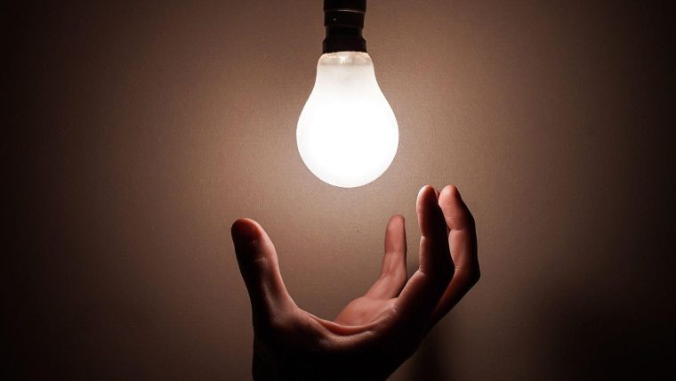 a hand reaching up to a bright lightbulb in a darkened room